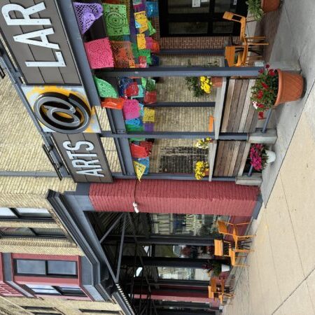 Street view of a brick building with a sign reading “ARTS @ LAR,” colorful paper banners, outdoor seating, and potted flowers by the entrance.