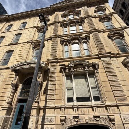 Tall, ornate stone building with large windows and decorative architectural details, viewed from street level on a sunny day. A streetlamp stands in front of the entrance.