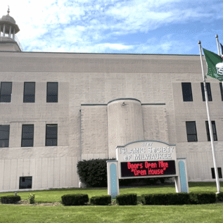 A beige mosque building with three flags, a dome, and a digital sign reading “Doors Open MKE ‘Open House’,” labeled “Islamic Society of Milwaukee.”.