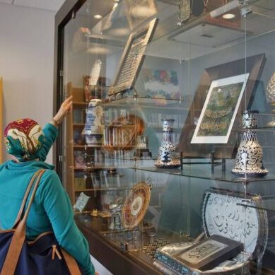 Two women wearing headscarves view intricate art and cultural artifacts displayed in a glass case at a museum or library exhibit.