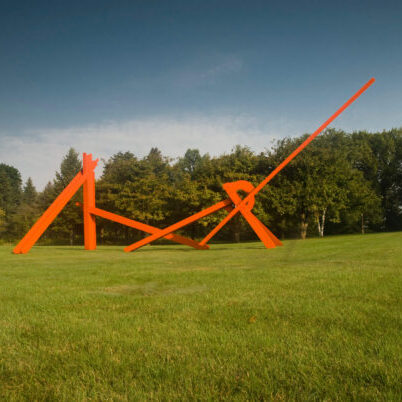 Large abstract orange metal sculpture with intersecting beams stands on a grassy field, with trees and blue sky in the background.