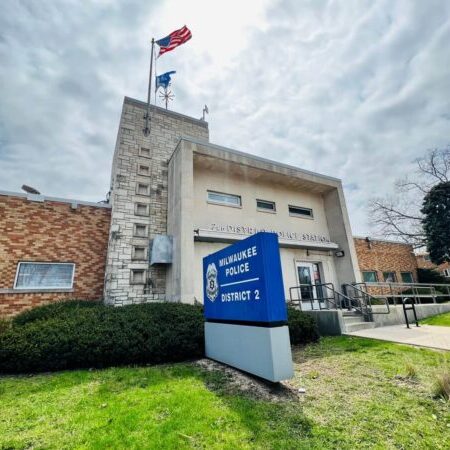 Exterior of Milwaukee Police District 2 station with signage, flagpoles, and a ramp leading to the entrance on a partly cloudy day.