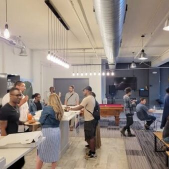 People gather in a modern office kitchen and lounge area, talking, eating, and playing pool. Some are seated at tables; others stand near the counter with pizza boxes visible.