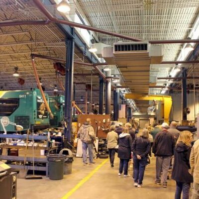 A group of people tours a large industrial facility with high ceilings, overhead lights, and buses being serviced.