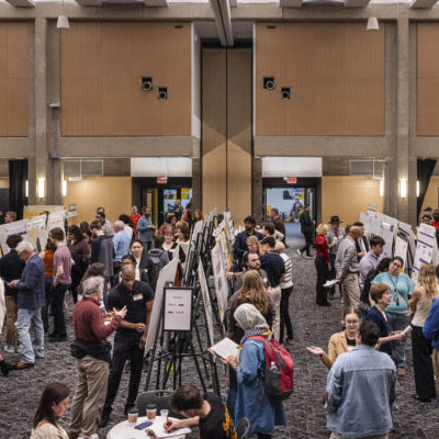 A large group of people view and discuss poster presentations at an indoor academic event in a spacious, well-lit hall.