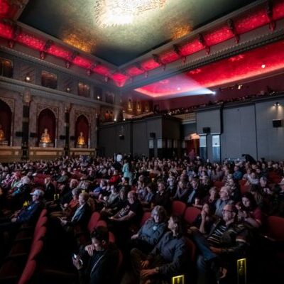 Audience seated in a dimly lit ornate theater with red lighting, watching an event or performance on stage.