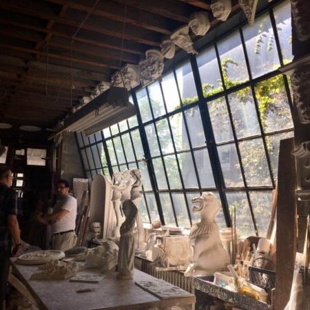 Two people talk in a sunlit workshop filled with sculpted plaster busts, statues, and classical architectural details near large slanted windows.