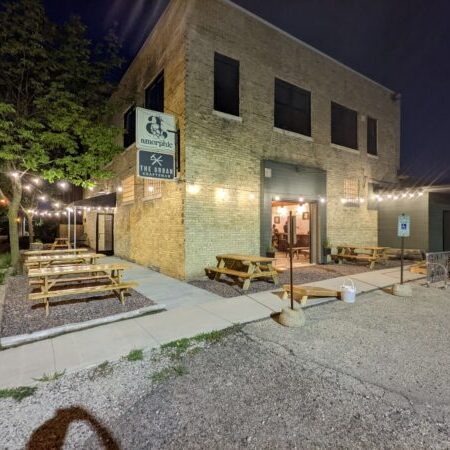 Outdoor seating area with picnic tables and string lights outside a brick building at night; several bicycles are parked near the entrance.
