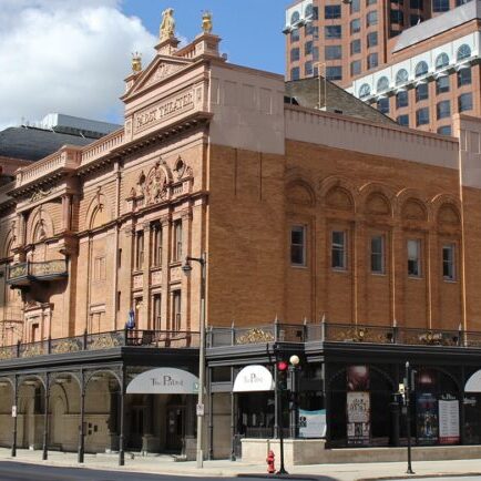 Historic theater building with ornate architecture, large arched windows, decorative statues on the roof, and a prominent “Pabst” sign on the corner; modern buildings in the background.