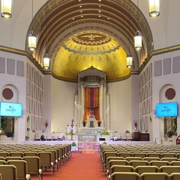 Interior of a church sanctuary with rows of beige chairs, a gold domed ceiling, large altar, colorful drapes, and two screens displaying event information.