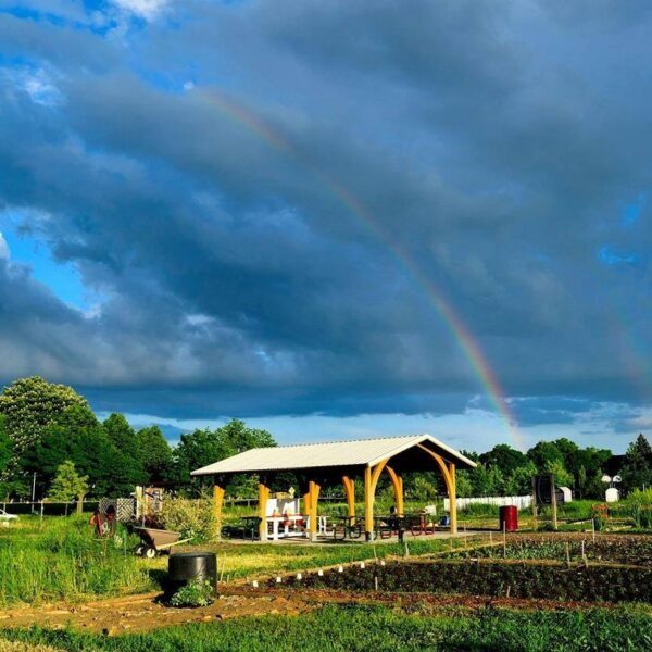 A rainbow arches over a wooden pavilion in a community garden under a cloudy sky, with trees and garden plots in the background.