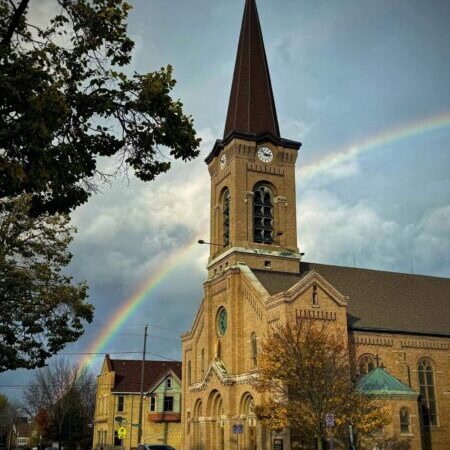 A brick church with a tall clock tower stands on a street corner under a cloudy sky, with a rainbow arching behind it and autumn trees nearby.