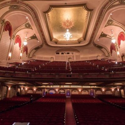 Elegant theater interior with ornate ceiling, red seats, balconies, and warm lighting, viewed from the stage facing the audience.
