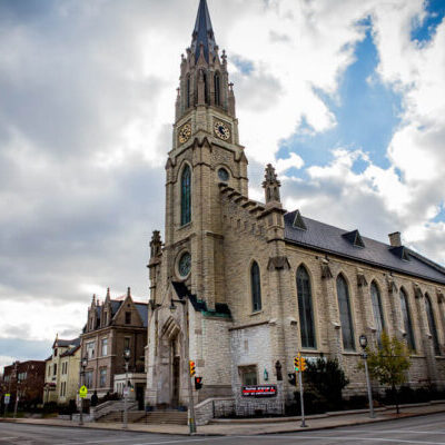 A large stone church with a tall clock tower stands at a street corner under a cloudy sky.