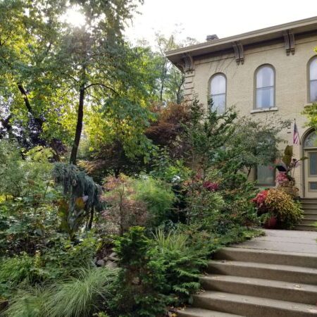 Two-story beige house with arched windows, surrounded by lush green plants and trees, with concrete steps leading to a porch decorated with potted plants.