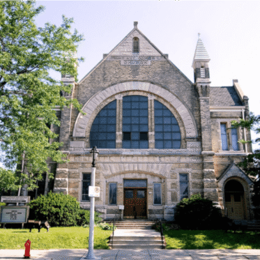 A large stone church with arched windows, twin steeples, and a central wooden door, surrounded by trees and a sidewalk with a fire hydrant in front.