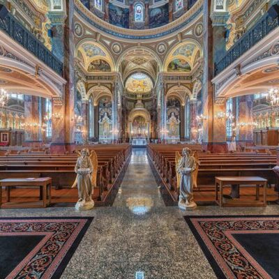 Ornate church interior with marble columns, wooden pews, statues, chandeliers, and detailed painted ceilings, viewed from the central aisle toward the altar.