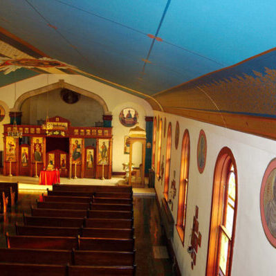 Interior of an Orthodox Christian church with wooden pews, iconostasis, religious icons, arched windows, and blue ceiling with gold accents.