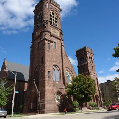 A large brown stone church with two towers stands on a street corner lined with trees, under a blue sky with scattered clouds. Several parked cars are visible along the street.