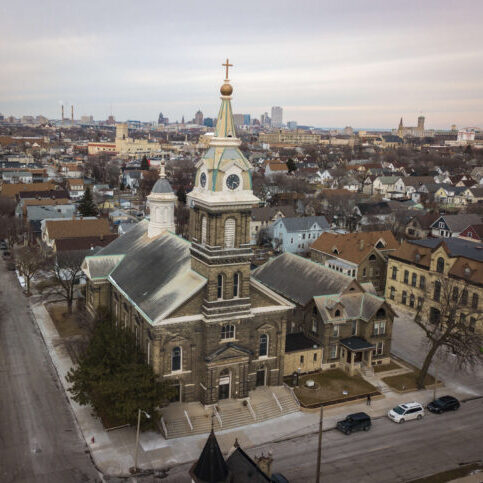 Aerial view of a large historic church with a tall clock tower in a residential neighborhood, city skyline visible in the background under cloudy skies.