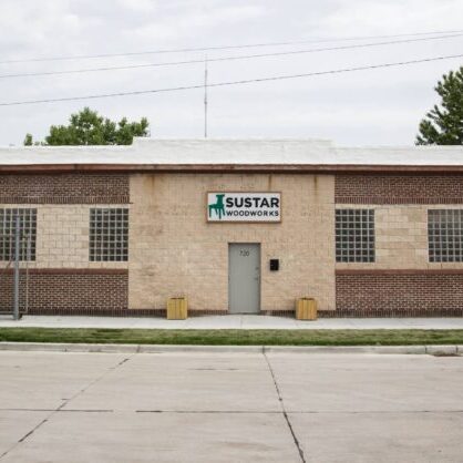 A single-story brick building with a sign reading “Sustar Woodworks” above the entrance, large windows, and two planters near the door.
