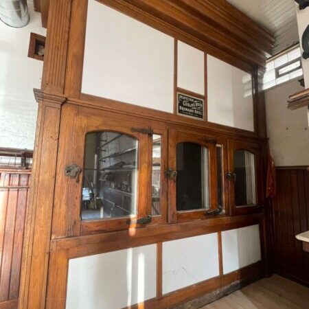 Vintage wooden service counter with glass windows and metal latches, set against white paneling; a sign above reads “Patrons must obtain checks from cashier before being served.”.