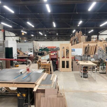 A spacious woodworking shop with various tools, machinery, workbenches, wood planks, and partially assembled doors, all organized in a well-lit industrial space.