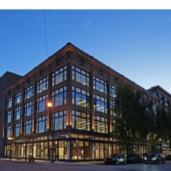 A four-story brick and glass building on a street corner at dusk, with large windows lit from inside and cars parked along the curb.