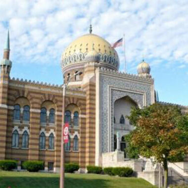 A large, ornate building with striped brickwork, arched windows, minarets, and a prominent golden dome under a partly cloudy sky. An American flag is visible on the roof.