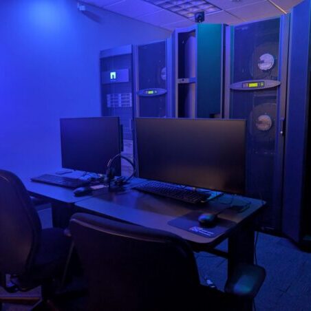 Two black office chairs and desks with monitors, keyboards, and headsets in front of tall server racks in a dimly lit, blue-toned room.