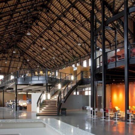 Spacious industrial-style office with exposed beams, a mezzanine, large windows, orange walls, open seating, desks, and a curved staircase.