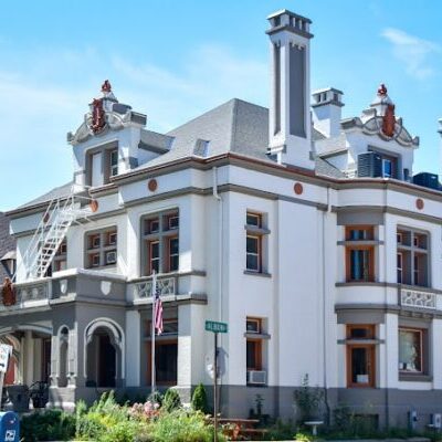 A large, historic two-story building with ornate architectural details, multiple chimneys, and a prominent front porch sits at a street corner on a sunny day.
