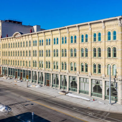 A four-story historic brick building with large arched windows on a sunny day, situated on a mostly empty city street with patches of snow on the sidewalks.