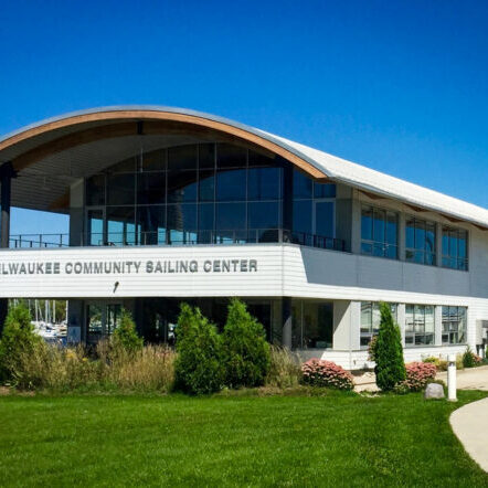 A modern two-story building with large windows labeled “Milwaukee Community Sailing Center,” surrounded by grass, shrubs, and trees under a clear blue sky.