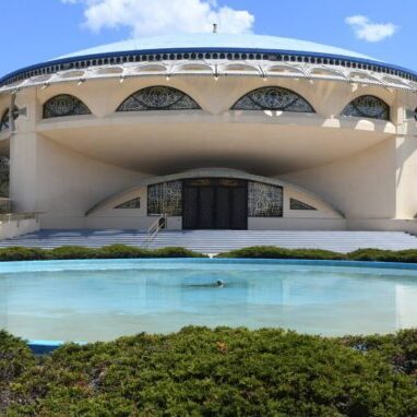 A round, modernist building with large arched windows and a dome roof stands behind a circular reflecting pool and landscaped bushes under a blue sky.