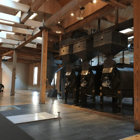 Industrial interior with wooden beams, skylights, and three large black roasting machines lined up on a tiled floor.