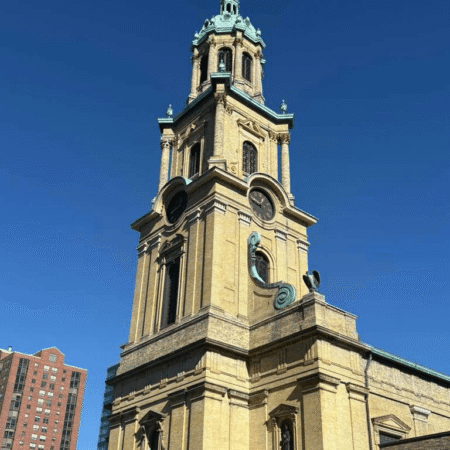 A tall, beige stone church with a prominent tower and green dome stands against a clear blue sky; a red brick building is seen in the background.