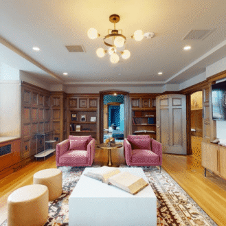 Library lounge with wood paneling, pink chairs, a rug, books, a large window, and a TV displaying “Journal Commons” on the wall.
