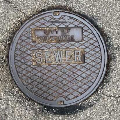 Round metal manhole cover labeled “City of Milwaukee SEWER” set into a textured asphalt street surface.