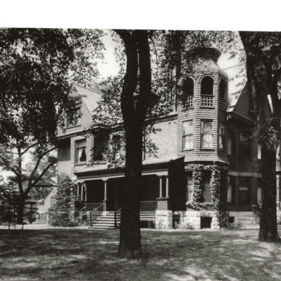Black-and-white photo of a large, ornate Victorian-style house with a prominent turret, wraparound porch, and trees in the front yard.