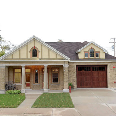 Two-story tan and brick house with a covered front porch, wooden front door, large garage door, and concrete driveway. There is a small lawn and a car visible at the side.