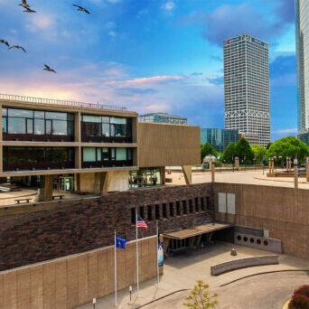 Modern urban plaza with a concrete building, flags, and a flock of birds flying overhead. Skyscrapers and trees are visible in the background under a blue sky with clouds.