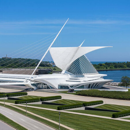 A modern white building with wing-like structures and cables sits by a body of water, surrounded by green lawns and trees under a clear blue sky.